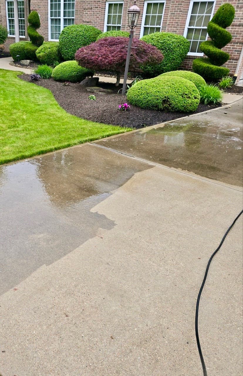 Driveway mid-clean: a bright freshly power-washed stripe against the untouched gray concrete, manicured shrubs and brick house behind